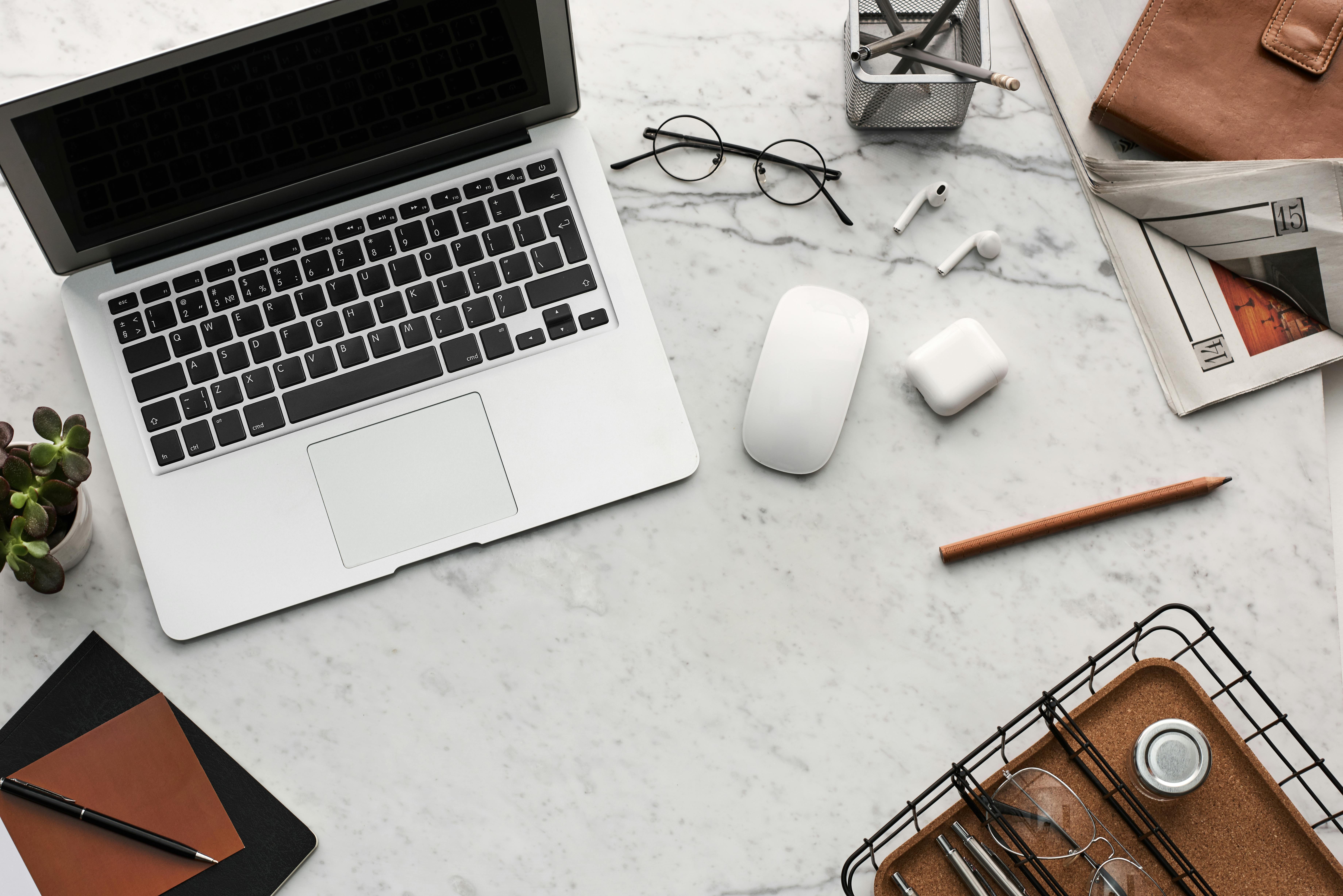 Top view of a workspace with laptop, mouse, glasses, earbuds, pencils, notebooks, and a plant on a marble desk.