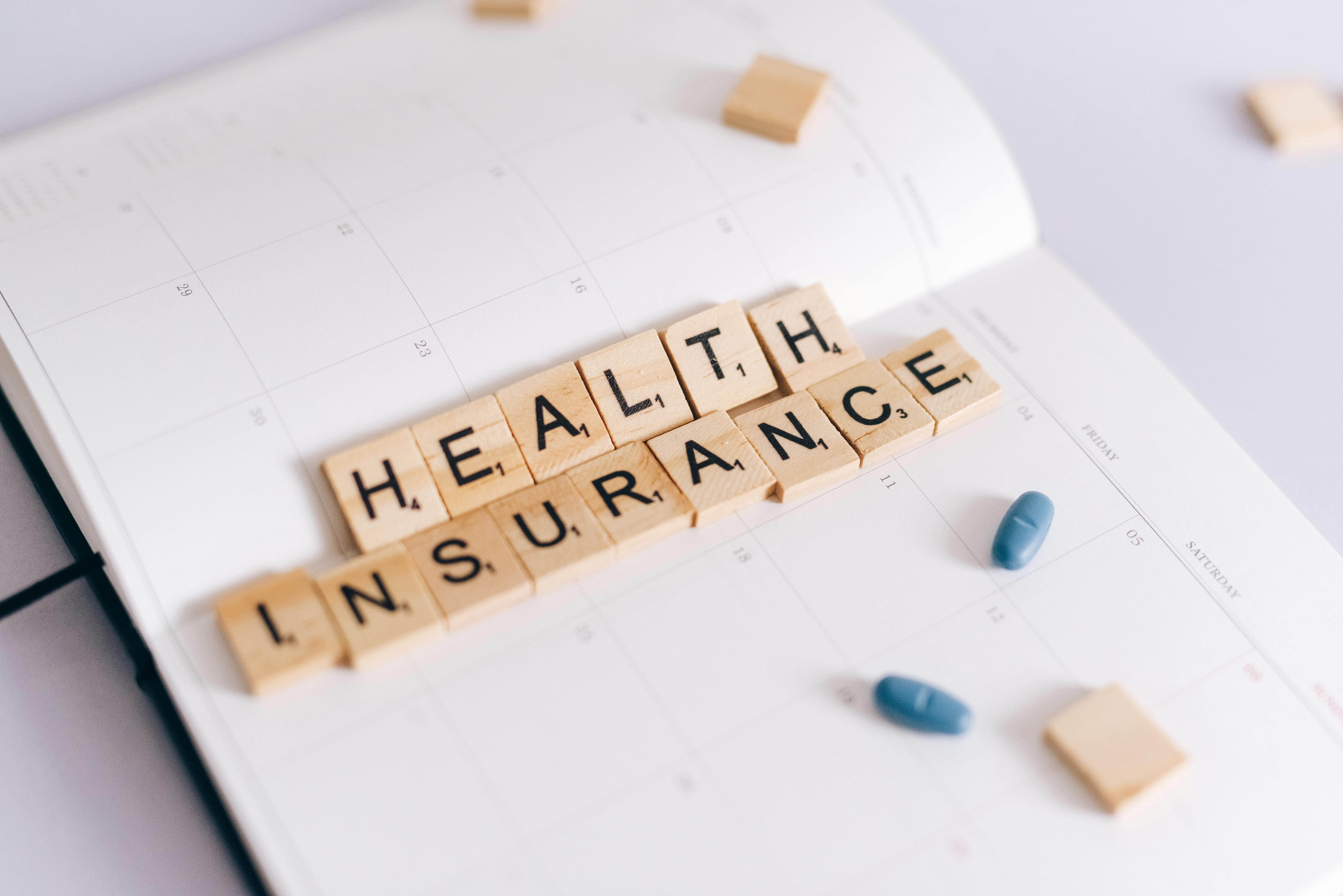 Scrabble tiles spelling health insurance on an open calendar with scattered blue pills.