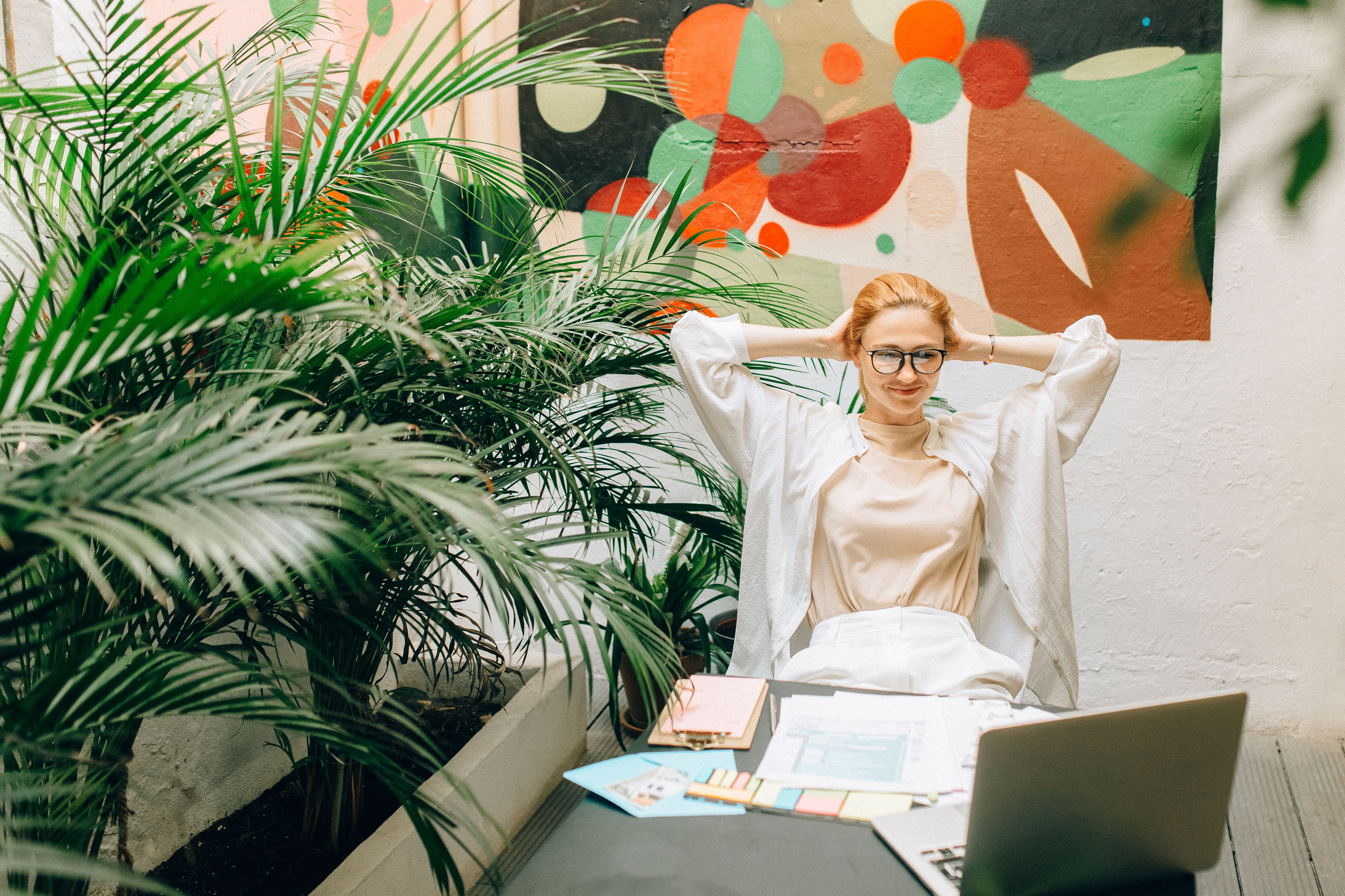 Woman with glasses relaxing with hands behind head at desk with laptop, papers, and plants nearby.
