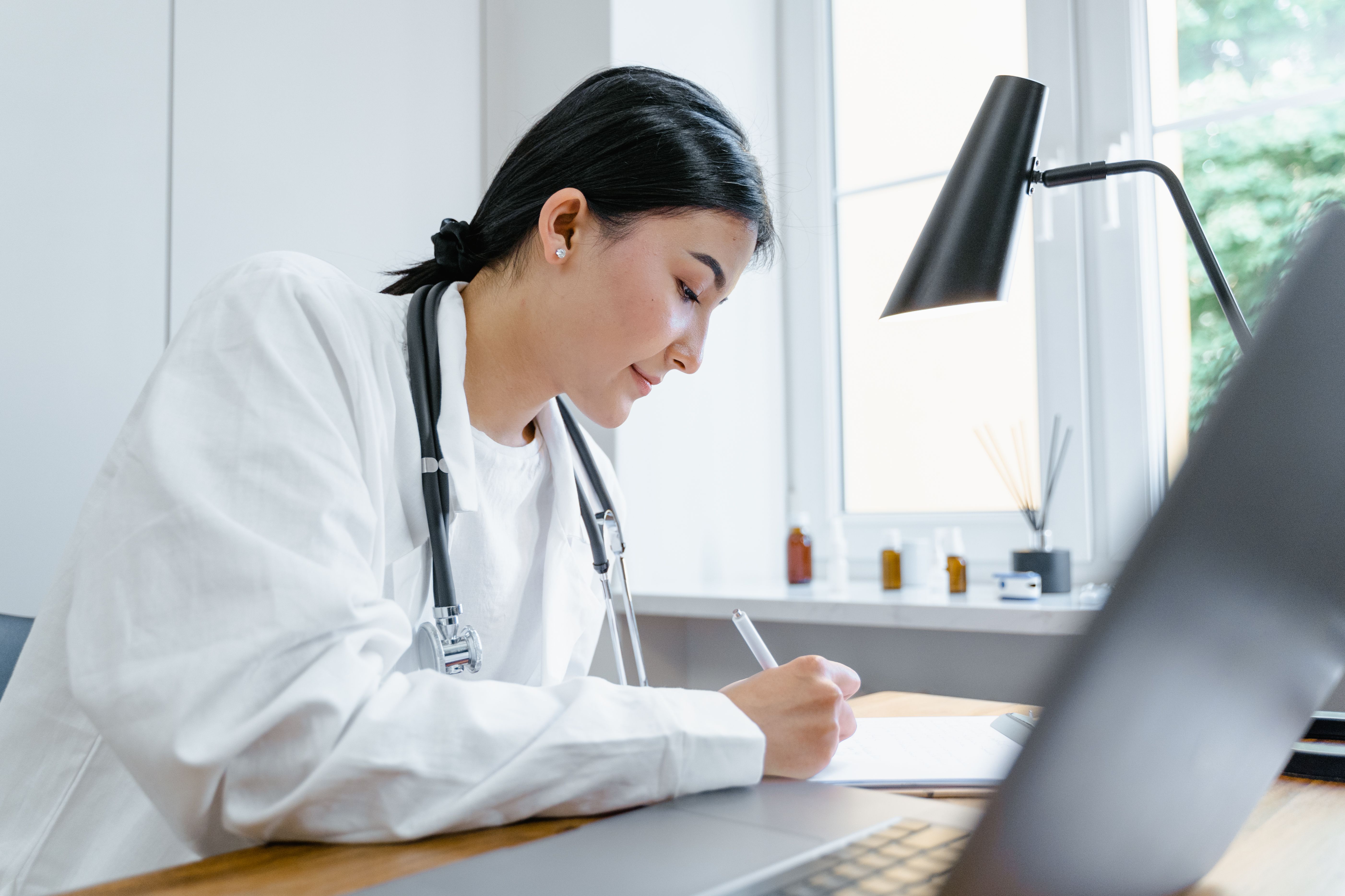 Female doctor with stethoscope writing notes at desk with laptop and lamp in bright office.